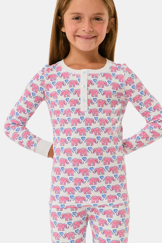 A young girl stands barefoot, smiling with hands on hips, wearing Kids Hathi Hearts Pajamas by Roller Rabbit—soft Pima Cotton pajamas featuring pink and blue elephant patterns on a white background.