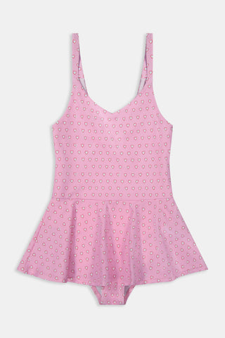 A young girl with long brown hair smiles as she wears the Roller Rabbit Kids Hearts Dory One Piece, a sleeveless pink dress with a flared, ruffled skirt and heart print, standing with her hands behind her back against a light background.