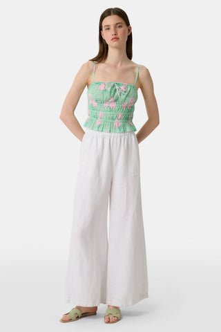 A woman wears the Roller Rabbit Hydrangea Gingham Aurelia Top—a green and white gingham crop top with pink hydrangea florals, thin straps, and a ruffled hem—paired with white pants against a plain white background.