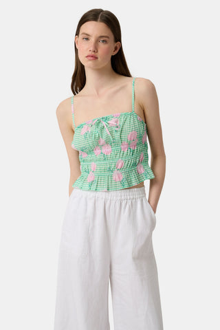 A woman wears the Roller Rabbit Hydrangea Gingham Aurelia Top—a green and white gingham crop top with pink hydrangea florals, thin straps, and a ruffled hem—paired with white pants against a plain white background.