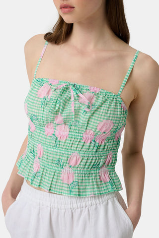 A woman wears the Roller Rabbit Hydrangea Gingham Aurelia Top—a green and white gingham crop top with pink hydrangea florals, thin straps, and a ruffled hem—paired with white pants against a plain white background.