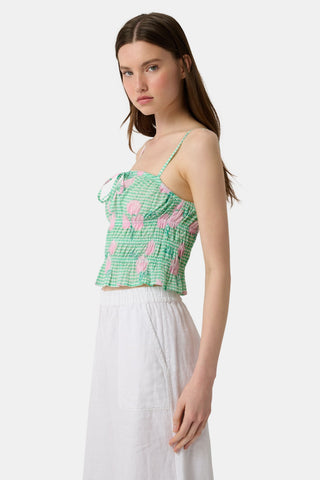 A woman wears the Roller Rabbit Hydrangea Gingham Aurelia Top—a green and white gingham crop top with pink hydrangea florals, thin straps, and a ruffled hem—paired with white pants against a plain white background.