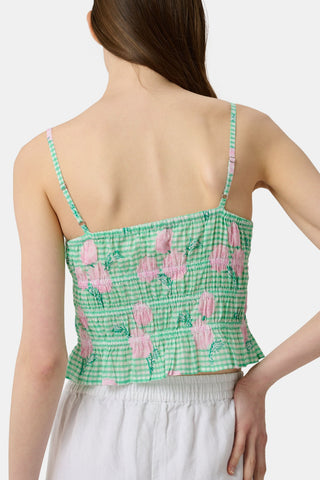 A woman wears the Roller Rabbit Hydrangea Gingham Aurelia Top—a green and white gingham crop top with pink hydrangea florals, thin straps, and a ruffled hem—paired with white pants against a plain white background.