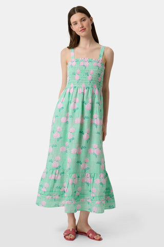 A woman wears the Roller Rabbit Hydrangea Gingham Bristol Dress, a long sleeveless style with pink florals, ruffled hem, and wide straps. She pairs it with pink sandals, standing with one hand by her side and the other on her leg.