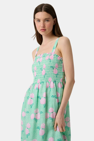 A woman wears the Roller Rabbit Hydrangea Gingham Bristol Dress, a long sleeveless style with pink florals, ruffled hem, and wide straps. She pairs it with pink sandals, standing with one hand by her side and the other on her leg.