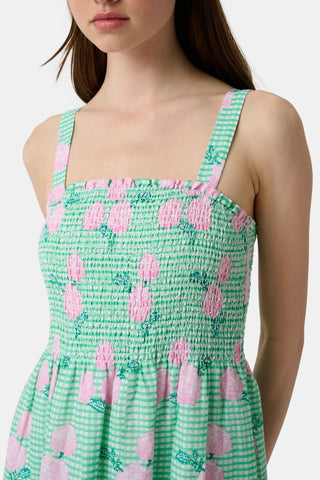 A woman wears the Roller Rabbit Hydrangea Gingham Bristol Dress, a long sleeveless style with pink florals, ruffled hem, and wide straps. She pairs it with pink sandals, standing with one hand by her side and the other on her leg.