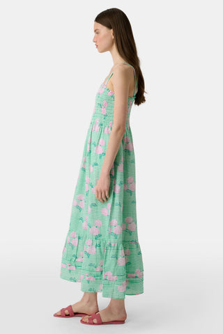 A woman wears the Roller Rabbit Hydrangea Gingham Bristol Dress, a long sleeveless style with pink florals, ruffled hem, and wide straps. She pairs it with pink sandals, standing with one hand by her side and the other on her leg.
