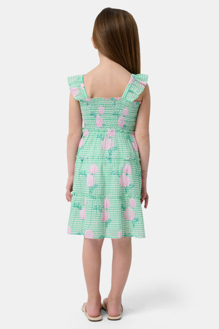 A young girl with straight brown hair smiles in the Roller Rabbit Kids Hydrangea Gingham Ceri Dress, featuring pink floral patterns and ruffled sleeves. She pairs it with white sandals and stands against a plain white background.