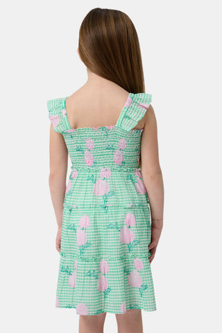 A young girl with straight brown hair smiles in the Roller Rabbit Kids Hydrangea Gingham Ceri Dress, featuring pink floral patterns and ruffled sleeves. She pairs it with white sandals and stands against a plain white background.