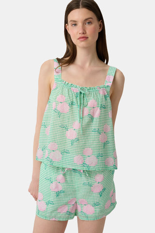 A woman with straight brown hair and a neutral expression wears the Roller Rabbit Hydrangea Gingham Cooper Set—green and white with pink floral prints. She stands with her hands in her shorts’ pockets and wears pink sandals.