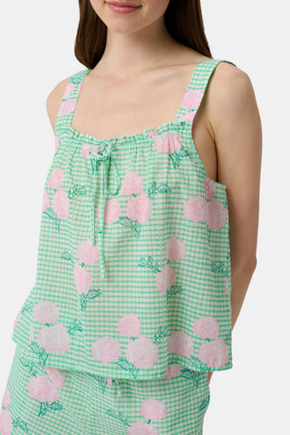 A woman with straight brown hair and a neutral expression wears the Roller Rabbit Hydrangea Gingham Cooper Set—green and white with pink floral prints. She stands with her hands in her shorts’ pockets and wears pink sandals.