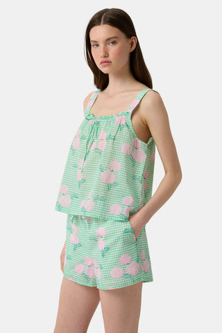 A woman with straight brown hair and a neutral expression wears the Roller Rabbit Hydrangea Gingham Cooper Set—green and white with pink floral prints. She stands with her hands in her shorts’ pockets and wears pink sandals.