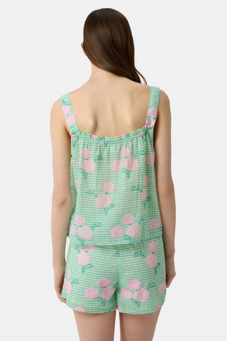 A woman with straight brown hair and a neutral expression wears the Roller Rabbit Hydrangea Gingham Cooper Set—green and white with pink floral prints. She stands with her hands in her shorts’ pockets and wears pink sandals.