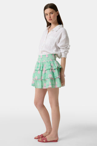 A person models the Roller Rabbit Hydrangea Gingham Siggy Skirt, featuring green and white checks with pink florals, paired with a tucked-in white shirt and red flat sandals. The image displays the skirt from waist to feet.