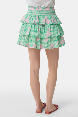 A person models the Roller Rabbit Hydrangea Gingham Siggy Skirt, featuring green and white checks with pink florals, paired with a tucked-in white shirt and red flat sandals. The image displays the skirt from waist to feet.