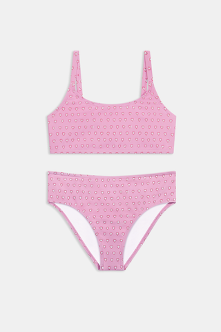 A teen girl stands smiling, wearing the Roller Rabbit Kids Hearts Lola Bikini—a light pink two-piece swimsuit with small heart patterns and full coverage bottoms—against a plain light gray background.
