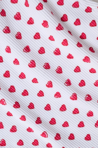 A child with long blonde hair wears the Roller Rabbit Kids Hearts Riley Set made from Pima Waffle fabric, featuring red hearts and ruffled cap sleeves, while holding a notebook with a decorative cover and standing in front of lockers.