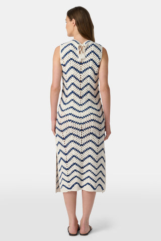 A woman wears the Roller Rabbit Zig Zag Irene Cover Up, a sleeveless, ankle-length crochet maxi dress with a blue and white zig zag pattern. She pairs it with black sandals and chunky striped bracelets, her straight brown hair worn loose.