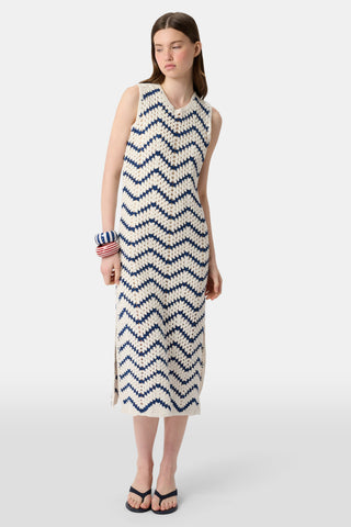 A woman wears the Roller Rabbit Zig Zag Irene Cover Up, a sleeveless crochet maxi dress with a blue and white zig zag pattern, paired with black sandals and large striped bangles, in front of a plain light background.