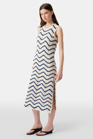 A woman wears the Roller Rabbit Zig Zag Irene Cover Up, a sleeveless crochet maxi dress with a blue and white zig zag pattern, paired with black sandals and large striped bangles, in front of a plain light background.