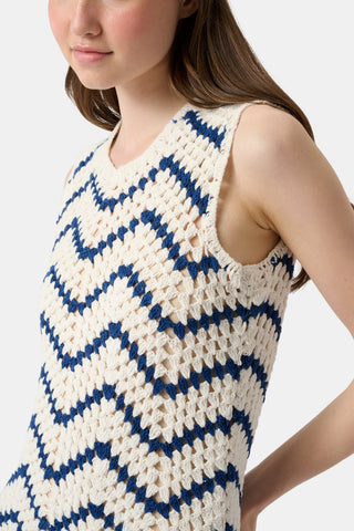 A woman wears the Roller Rabbit Zig Zag Irene Cover Up, a sleeveless crochet maxi dress with a blue and white zig zag pattern, paired with black sandals and large striped bangles, in front of a plain light background.