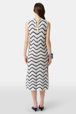 A woman wears the Roller Rabbit Zig Zag Irene Cover Up, a sleeveless crochet maxi dress with a blue and white zig zag pattern, paired with black sandals and large striped bangles, in front of a plain light background.