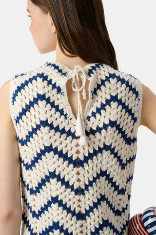 A woman wears the Roller Rabbit Zig Zag Irene Cover Up, a sleeveless crochet maxi dress with a blue and white zig zag pattern, paired with black sandals and large striped bangles, in front of a plain light background.