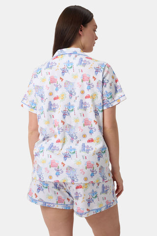 A woman with long dark hair wears Roller Rabbit’s Italy Monkey Polo Pajamas, a playful Pima Cotton set featuring cartoon animals and Italian motifs. The short-sleeved, button-up top pairs perfectly with the matching shorts.