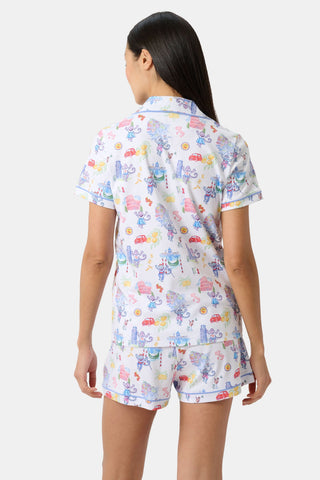 A woman with long dark hair wears Roller Rabbit’s Italy Monkey Polo Pajamas, a playful Pima Cotton set featuring cartoon animals and Italian motifs. The short-sleeved, button-up top pairs perfectly with the matching shorts.