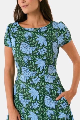 A woman wearing a SAPPHIRE Antonia Floral Jennifer Dress with a green and blue floral print, cap sleeves, tiered midi skirt, and side pockets, paired with black heeled sandals.