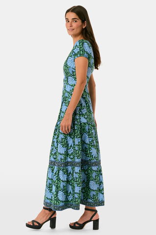 A woman wearing a SAPPHIRE Antonia Floral Jennifer Dress with a green and blue floral print, cap sleeves, tiered midi skirt, and side pockets, paired with black heeled sandals.