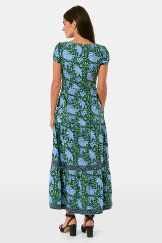 A woman wearing a SAPPHIRE Antonia Floral Jennifer Dress with a green and blue floral print, cap sleeves, tiered midi skirt, and side pockets, paired with black heeled sandals.