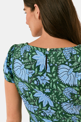 A woman wearing a SAPPHIRE Antonia Floral Jennifer Dress with a green and blue floral print, cap sleeves, tiered midi skirt, and side pockets, paired with black heeled sandals.