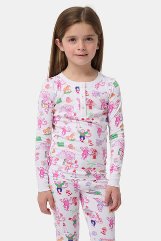 Young girl with straight, light brown hair wears white pajamas decorated with colorful illustrations of animals, food, and playful objects, standing against a plain light gray background.
