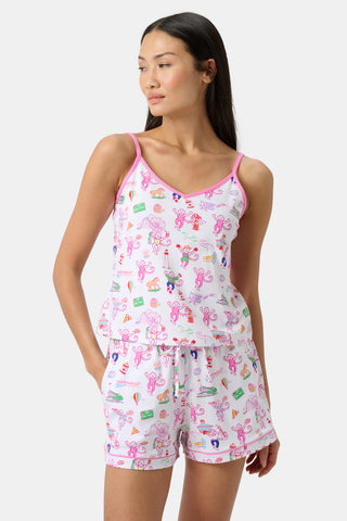 Woman wearing white pajama set with pink trim and playful cartoon prints, including bunnies and colorful objects. She stands with one hand in her pocket, looking to the side, for a charming touch against a plain background.