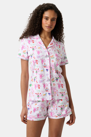 A woman with curly hair wears a matching short-sleeve pajama set with a white base and colorful cartoon print, standing against a plain light grey background. The playful design of her pajamas adds a cheerful touch to her look.