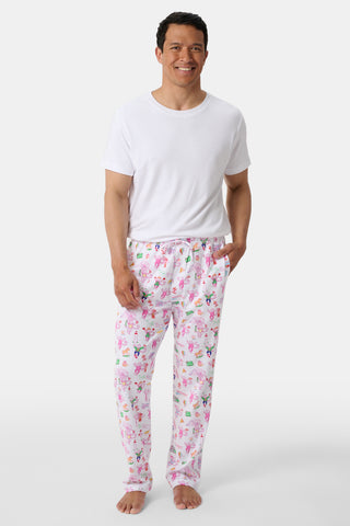 A man stands barefoot wearing a white t-shirt and colorful pajama pants with playful patterns, smiling at the camera against a plain white backdrop.