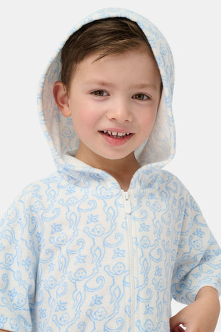 A young child smiles at the camera while wearing the Roller Rabbit Kids Monkey Jesse Cover Up, a light blue and white terry zip-up onesie with a playful animal pattern, against a plain background.