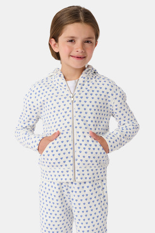 A young girl with curly hair wears the Kids Hearts Harley Hoodie, a cozy blue heart print french terry pajama set, standing against a plain light background with one hand in her pocket.