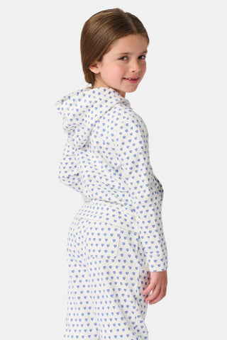 A young girl with curly hair wears the Kids Hearts Harley Hoodie, a cozy blue heart print french terry pajama set, standing against a plain light background with one hand in her pocket.