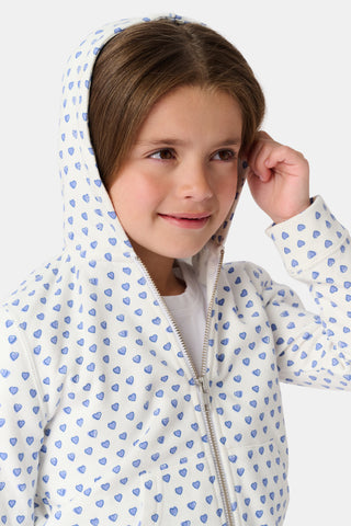 A young girl with curly hair wears the Kids Hearts Harley Hoodie, a cozy blue heart print french terry pajama set, standing against a plain light background with one hand in her pocket.