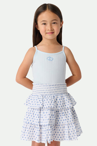 A young girl with long brown hair smiles, wearing the Roller Rabbit Kids Hearts Pointelle Bridget Cami with spaghetti straps and a tiered white skirt with blue hearts, hands behind her back, against a plain light background.