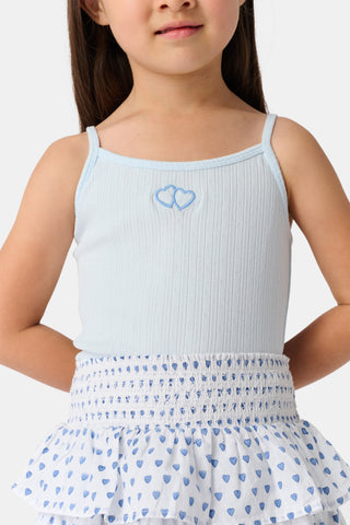 A young girl with long brown hair smiles, wearing the Roller Rabbit Kids Hearts Pointelle Bridget Cami with spaghetti straps and a tiered white skirt with blue hearts, hands behind her back, against a plain light background.