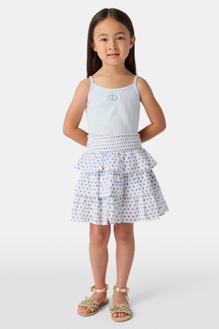 A young girl with long brown hair smiles, wearing the Roller Rabbit Kids Hearts Pointelle Bridget Cami with spaghetti straps and a tiered white skirt with blue hearts, hands behind her back, against a plain light background.