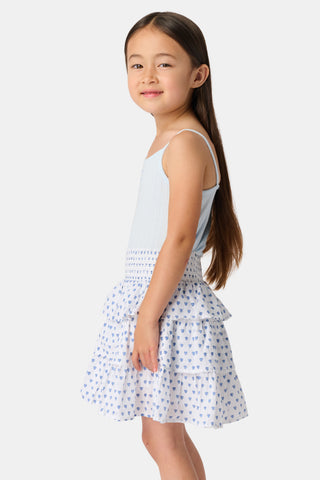 A young girl with long brown hair smiles, wearing the Roller Rabbit Kids Hearts Pointelle Bridget Cami with spaghetti straps and a tiered white skirt with blue hearts, hands behind her back, against a plain light background.