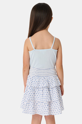 A young girl with long brown hair smiles, wearing the Roller Rabbit Kids Hearts Pointelle Bridget Cami with spaghetti straps and a tiered white skirt with blue hearts, hands behind her back, against a plain light background.