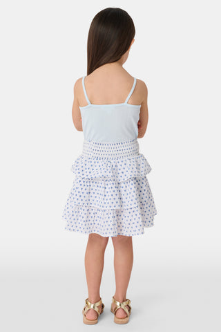 A young girl with long brown hair smiles, wearing the Roller Rabbit Kids Hearts Pointelle Bridget Cami with spaghetti straps and a tiered white skirt with blue hearts, hands behind her back, against a plain light background.