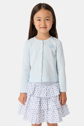 A young woman with straight brown hair stands outdoors near greenery, looking down while wearing a Roller Rabbit Kids Hearts Pointelle Heidi Cardigan and matching embroidered top, holding her cardigan open slightly with one hand.