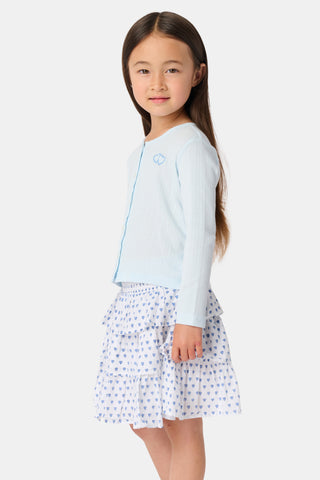 A young girl with long brown hair wears a light blue Roller Rabbit Kids Hearts Pointelle Heidi Cardigan over a white tiered skirt with blue heart patterns, standing and smiling against a plain white background.