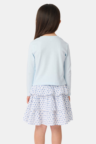 A young girl with long brown hair wears a light blue Roller Rabbit Kids Hearts Pointelle Heidi Cardigan over a white tiered skirt with blue heart patterns, standing and smiling against a plain white background.